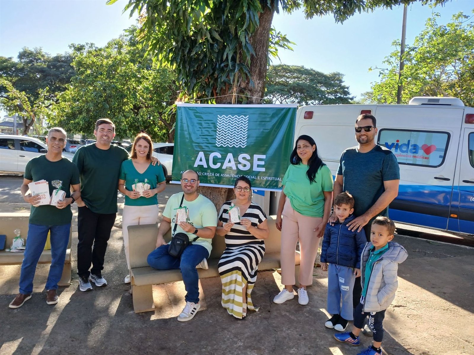 Voluntários e famílias seguram livros diante da faixa verde da ACASE, ao lado de uma ambulância.