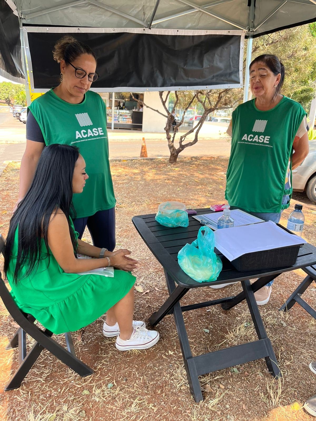 Voluntárias da ACASE oram com uma mulher sentada à mesa da tenda.