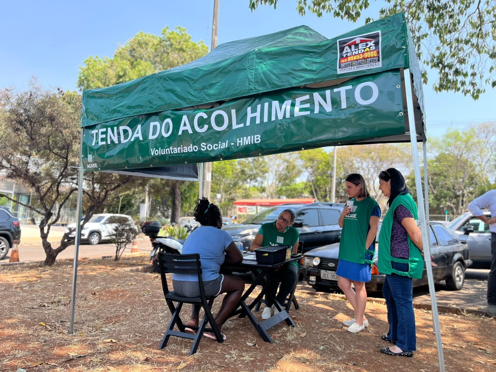 Voluntárias da ACASE atendem uma mãe à mesa, sob a tenda verde escrita “Tenda do Acolhimento – Voluntariado Social – HMIB”.
