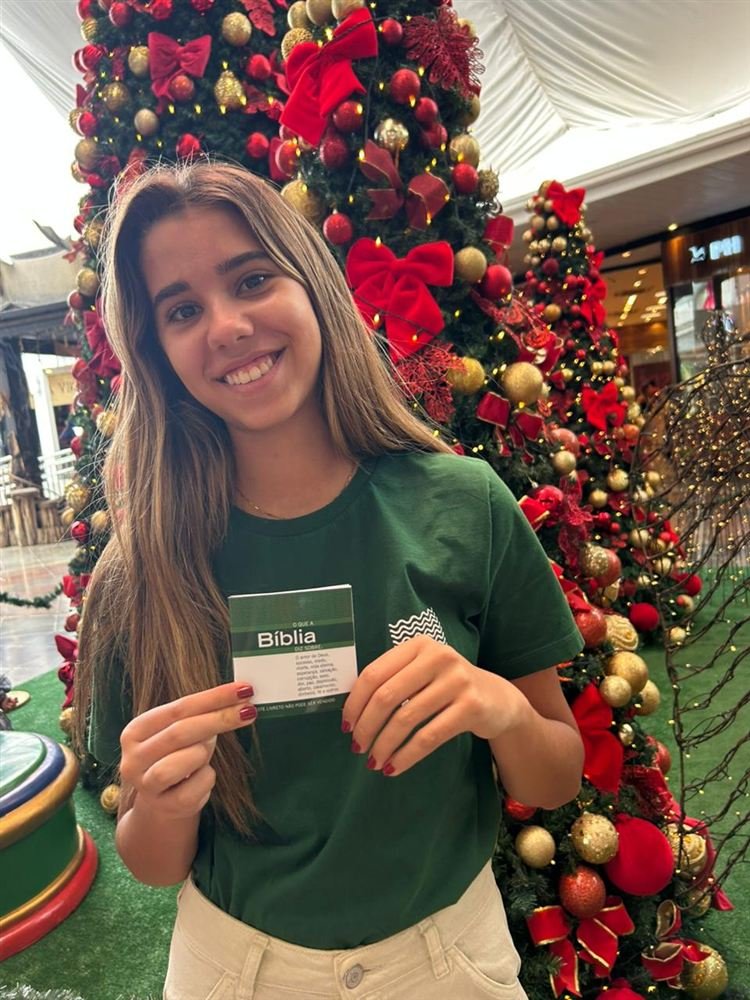Voluntária teen da ACASE mostra uma mini Bíblia diante de uma árvore de Natal.